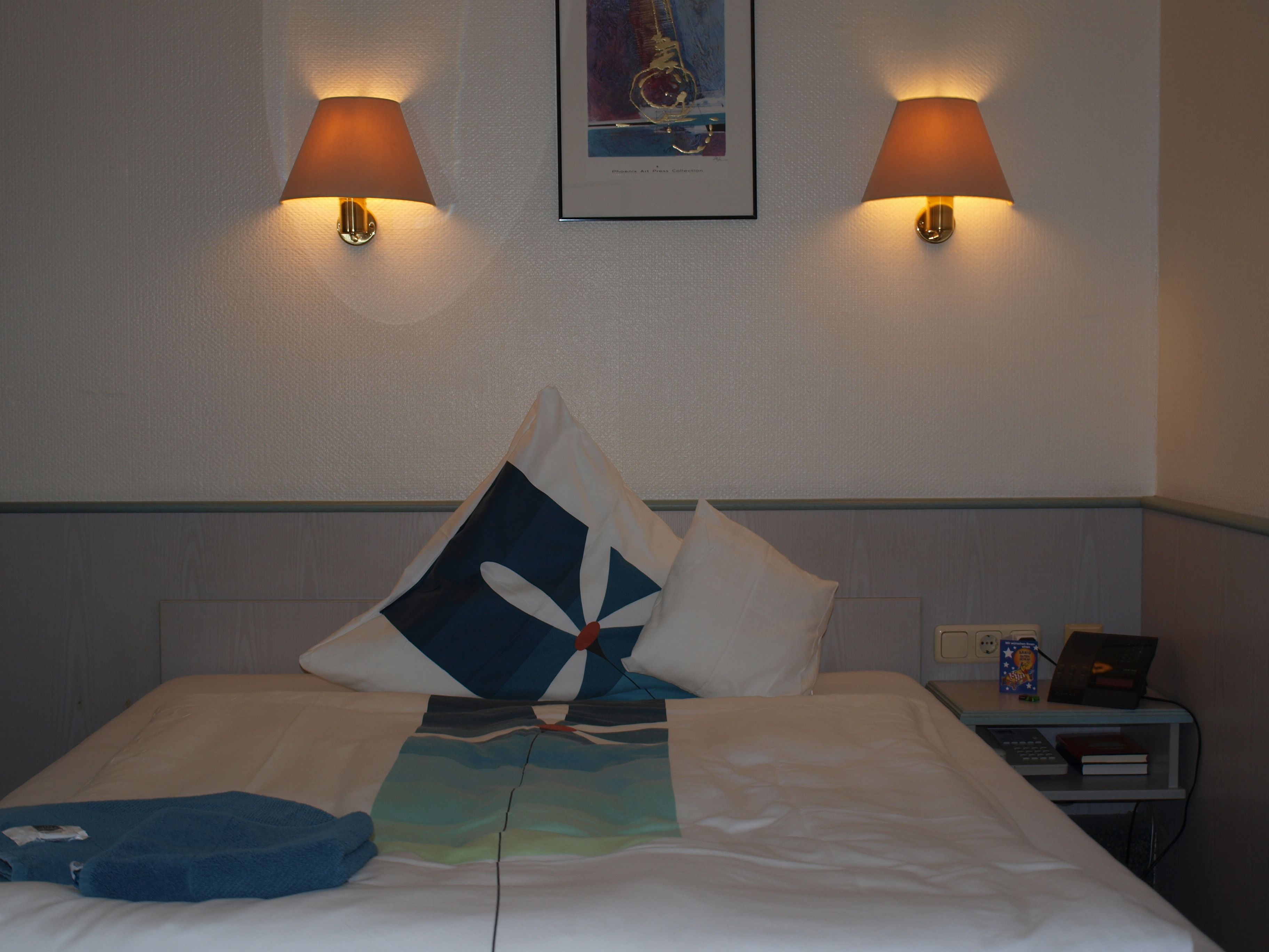 Una habitación de hotel con una cama bien hecha, dos lámparas de pared, una obra enmarcada sobre la cama y una mesita de noche con un teléfono y un libro.