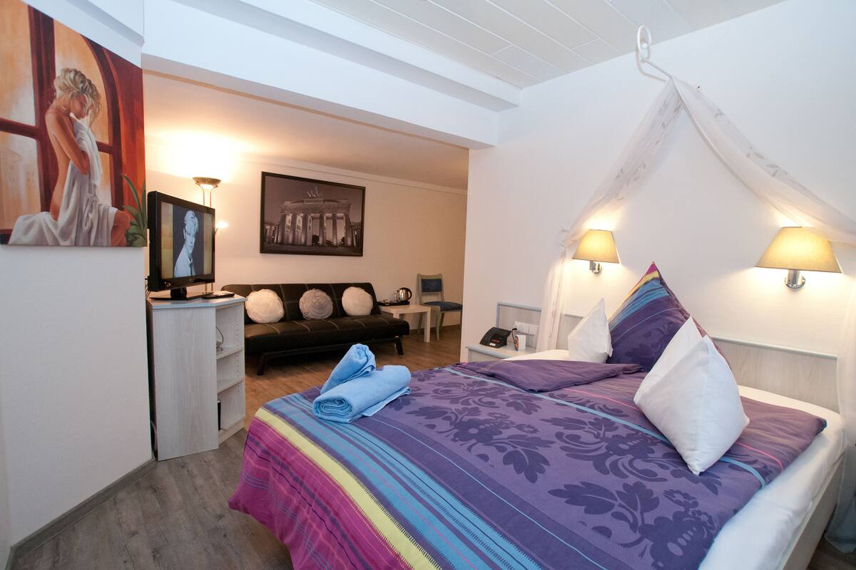 A bedroom with a canopy bed, a desk, a chair, a sofa, and artwork on the walls.