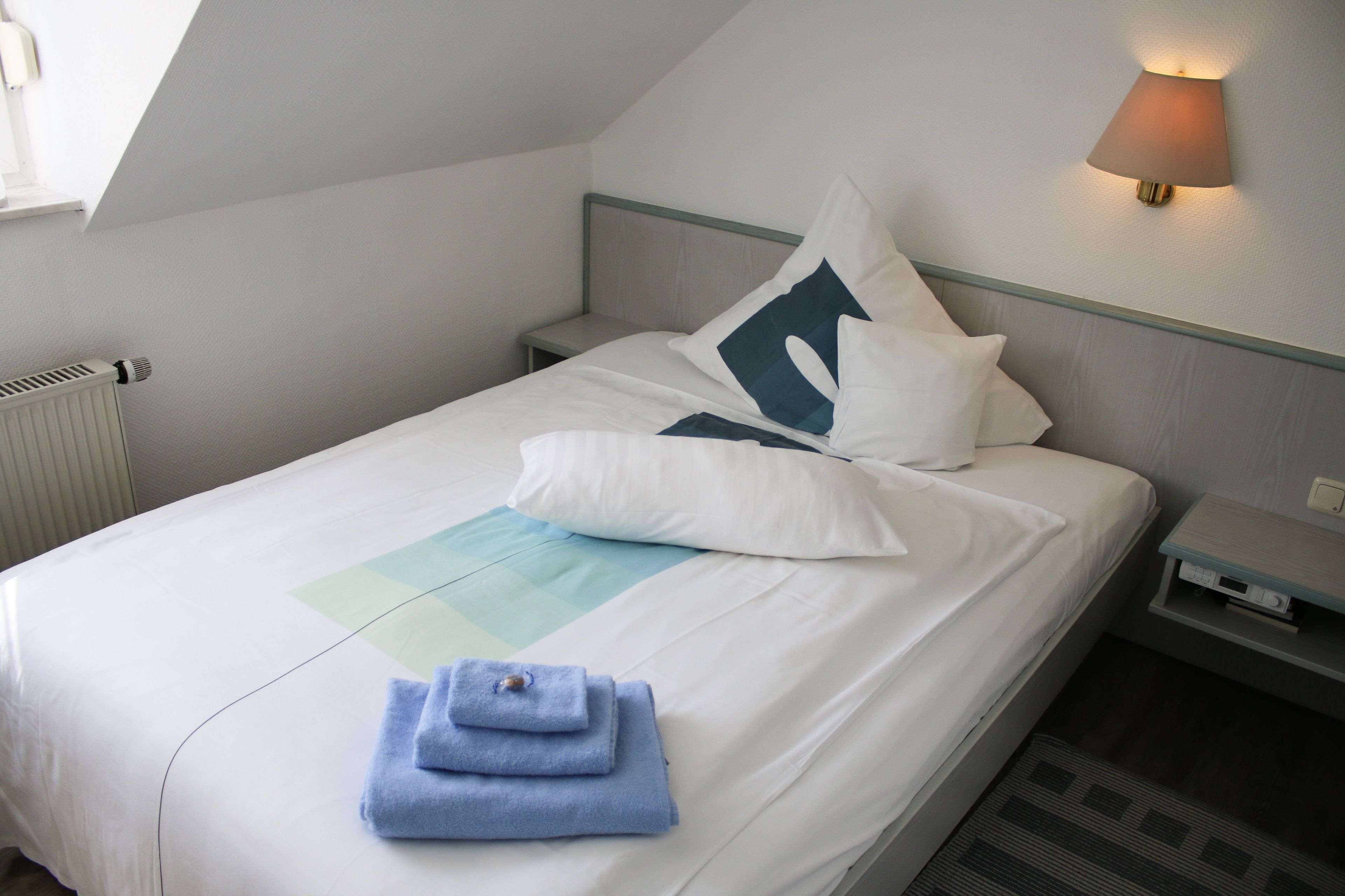 A neatly made bed with white linens, a blue towel, and a geometric-patterned pillow.
