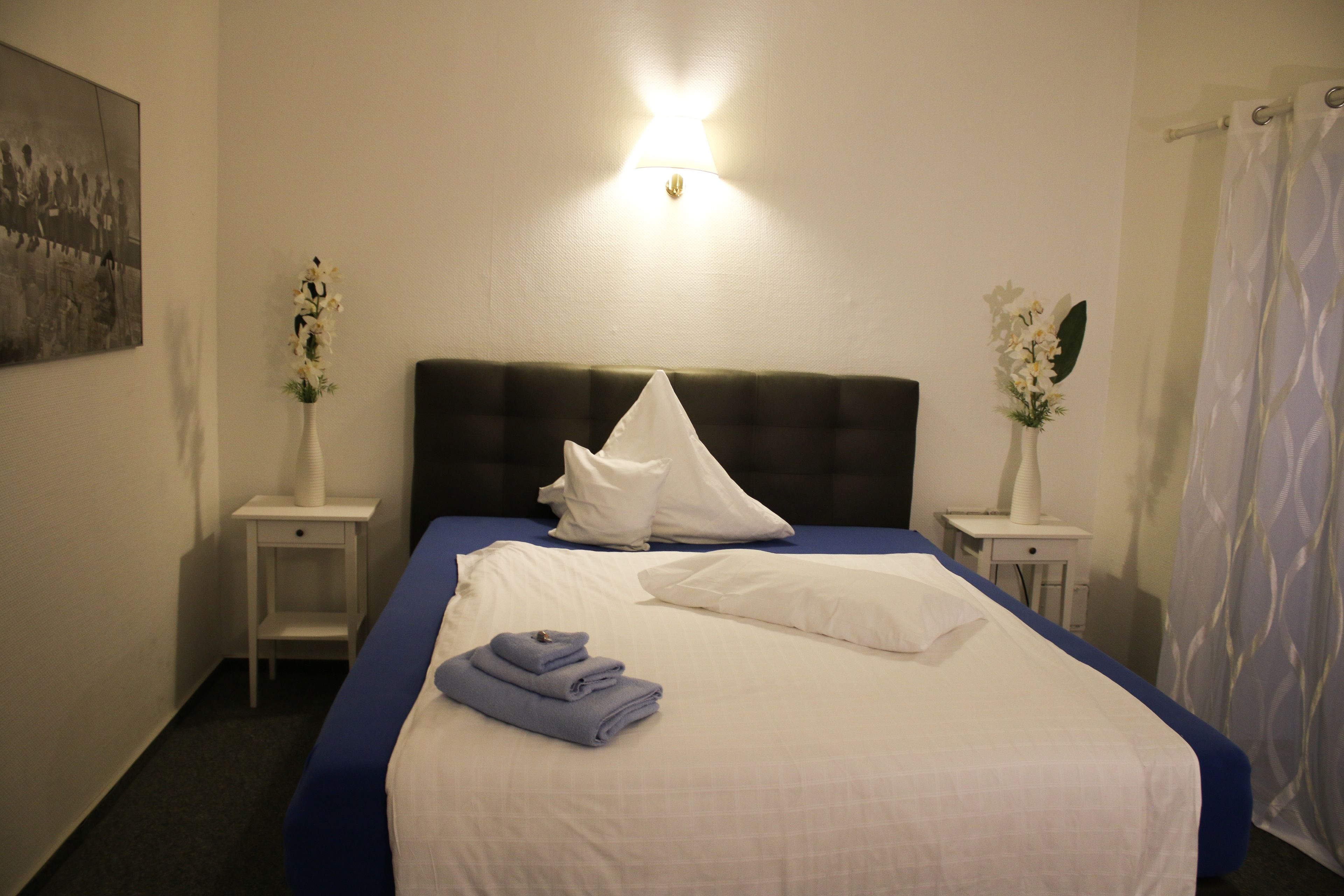 Un lit bien fait, avec une literie blanche et bleue, deux tables de chevet avec des lampes et un luminaire fixé au mur.
