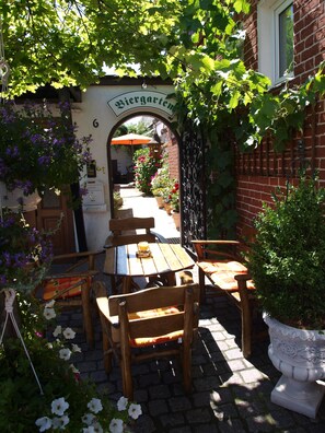 Un bâtiment en briques, doté d’une arche couverte de lierre sur laquelle on peut lire l’inscription « Biergarten », qui mène à un espace extérieur aménagé avec des tables et des chaises en bois.