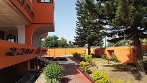 A courtyard with orange walls, a covered walkway, and a variety of trees and shrubs. - Tribeca Hotel Annex (Koforidua)