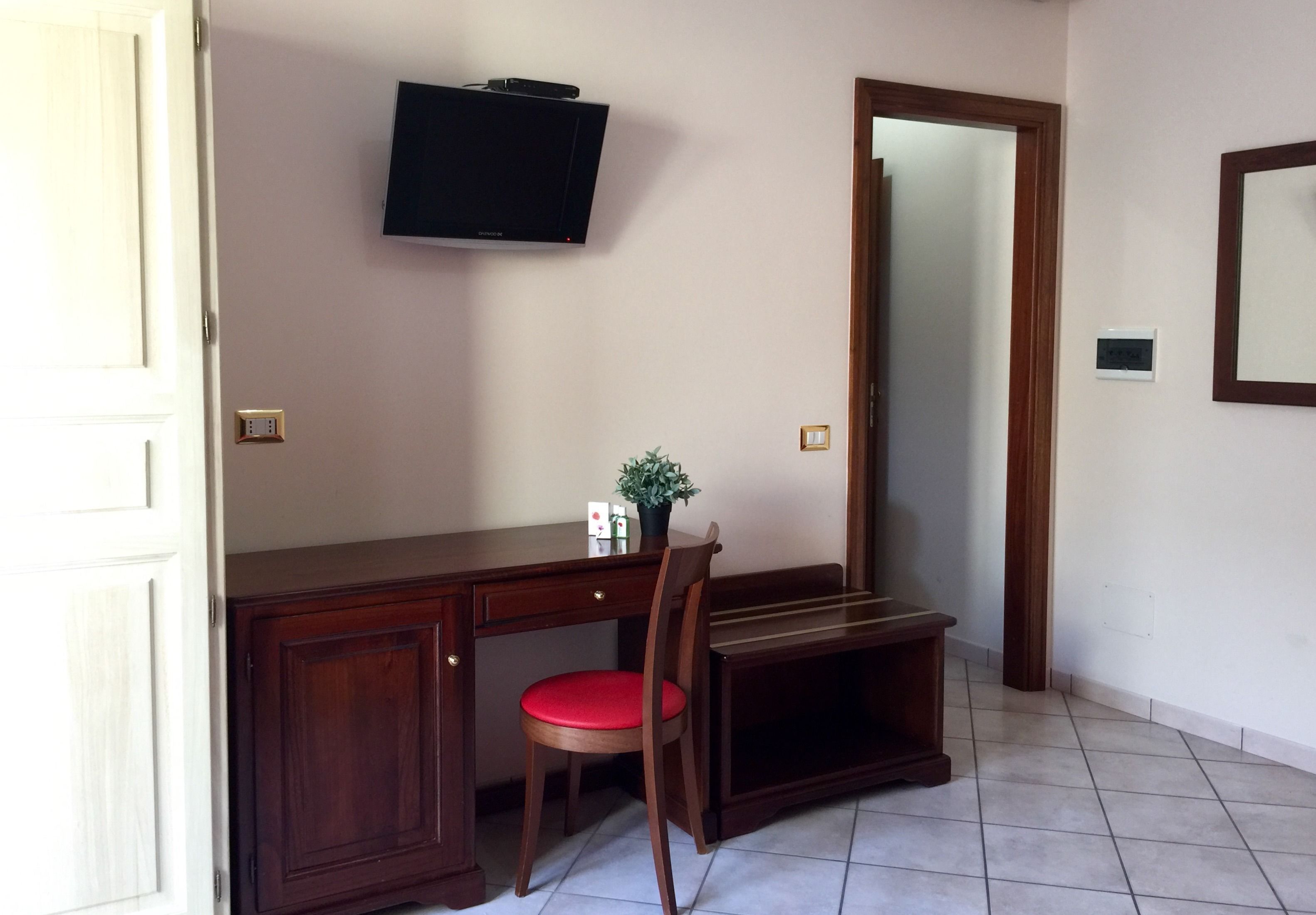A room with a wooden desk, a red cushioned chair, a television mounted on the wall, and a mirror on the wall.