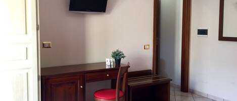 A room with a wooden desk, a red cushioned chair, a television mounted on the wall, and a mirror on the wall.