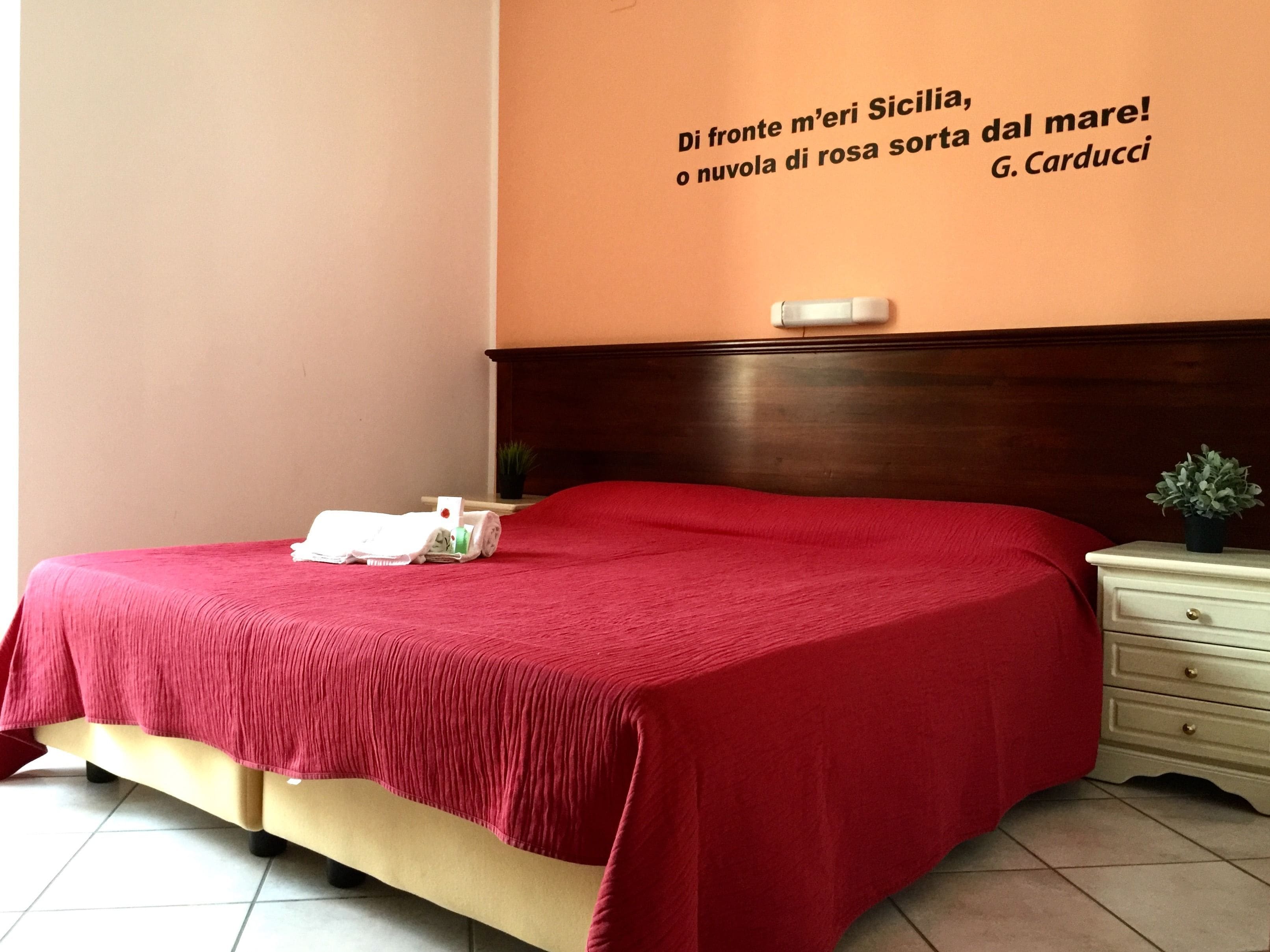 A hotel room with a red bedspread, a wooden headboard, a bedside table with a plant, and a wall with a quote in Italian.