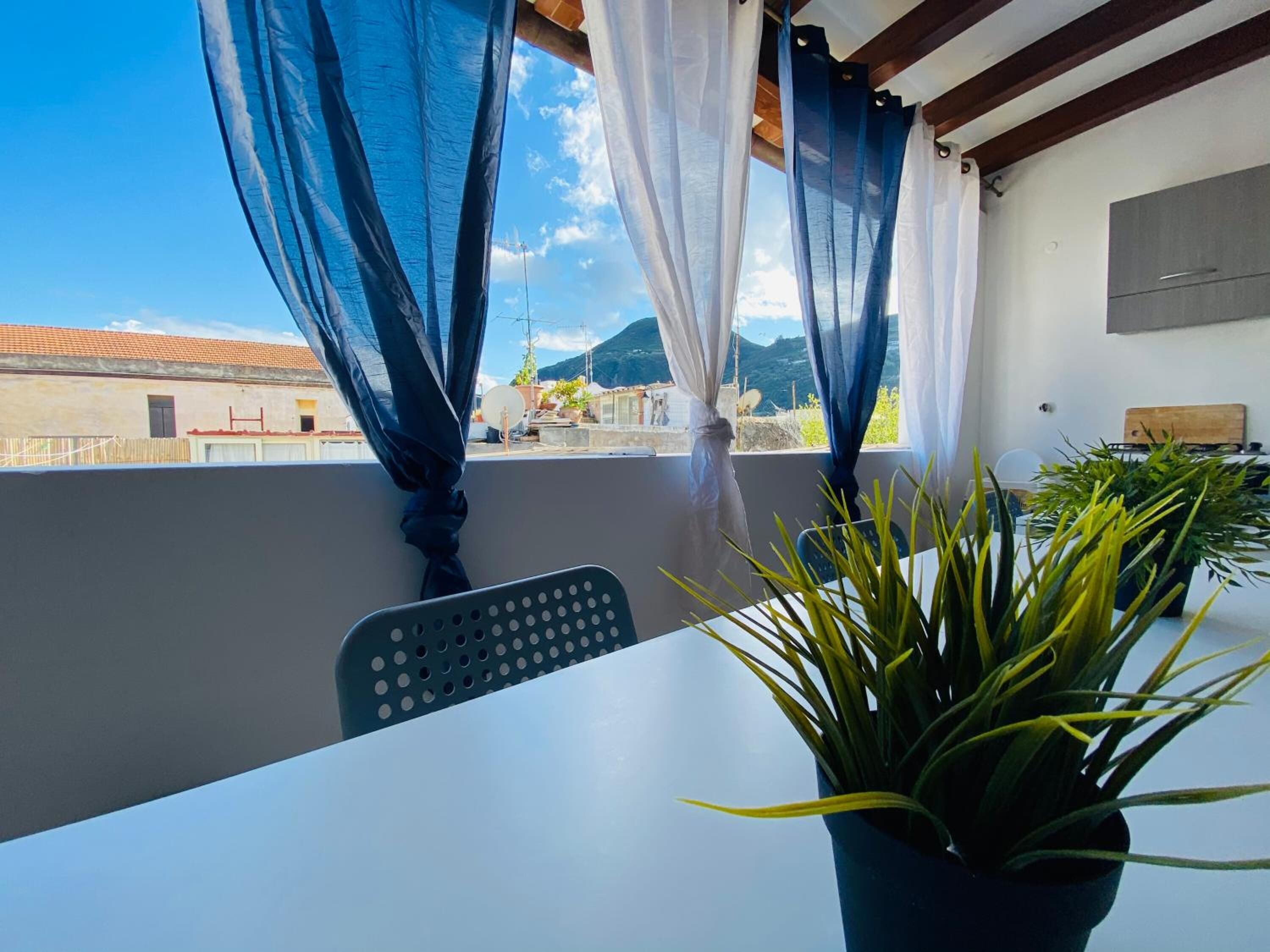 A balcony with a table and chair, a potted plant, and a view of buildings and mountains.