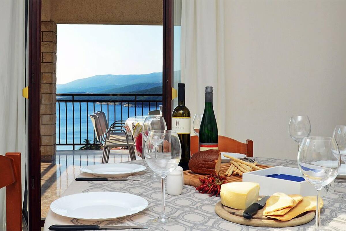 Una mesa de comedor dispuesta con platos, cubiertos, copas de vino y alimentos, con vistas a un paisaje con montañas y una masa de agua.