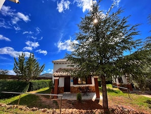 A building with a tiled roof, a small porch with chairs, and a tree in the foreground. - Complejo Rural Ibipozo (Pozo Alcon)