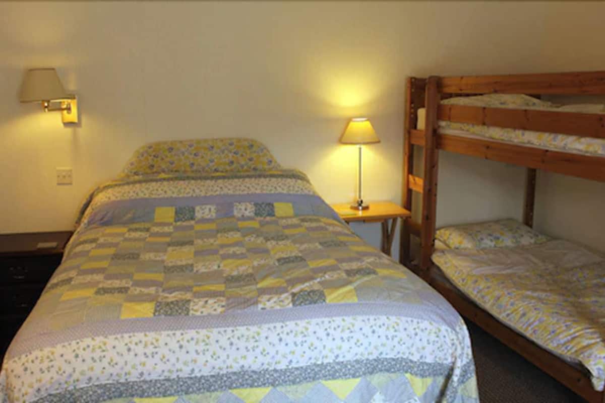 Una habitación con una cama grande, una cama litera más pequeña, una mesita de noche con lámpara y un aplique en la pared.