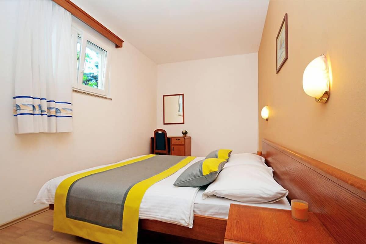 Ein Schlafzimmer mit Bett, Nachttisch, Fenster mit Vorhängen, Wandlampe und einem kleinen Spiegel.