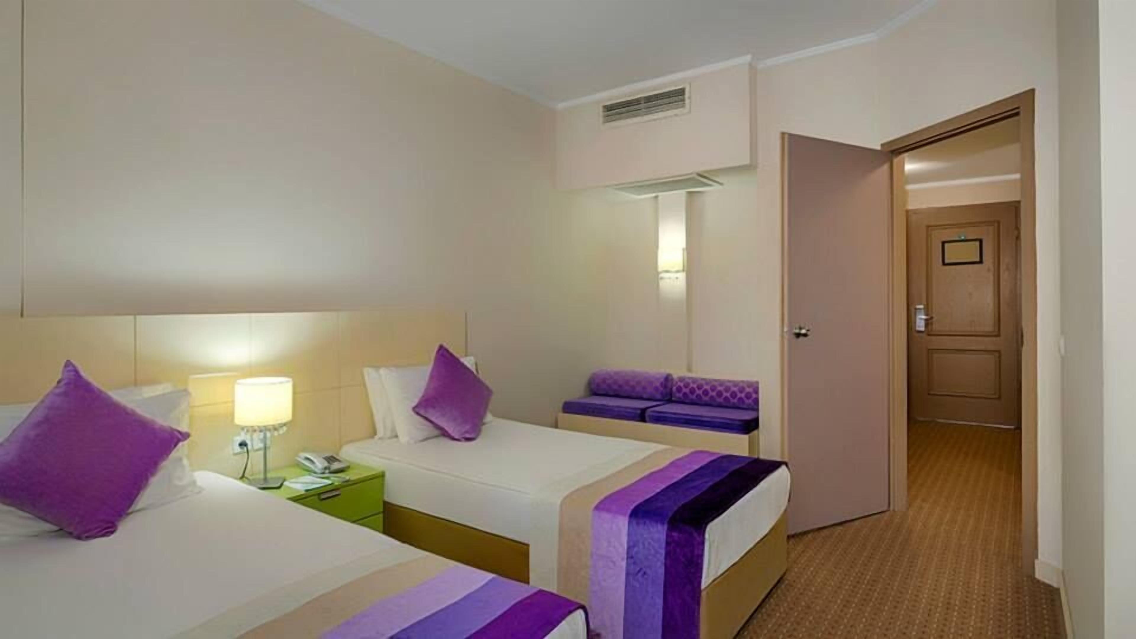 A hotel room with two beds, a bedside table with a lamp, a small seating area, and a door leading to another room.