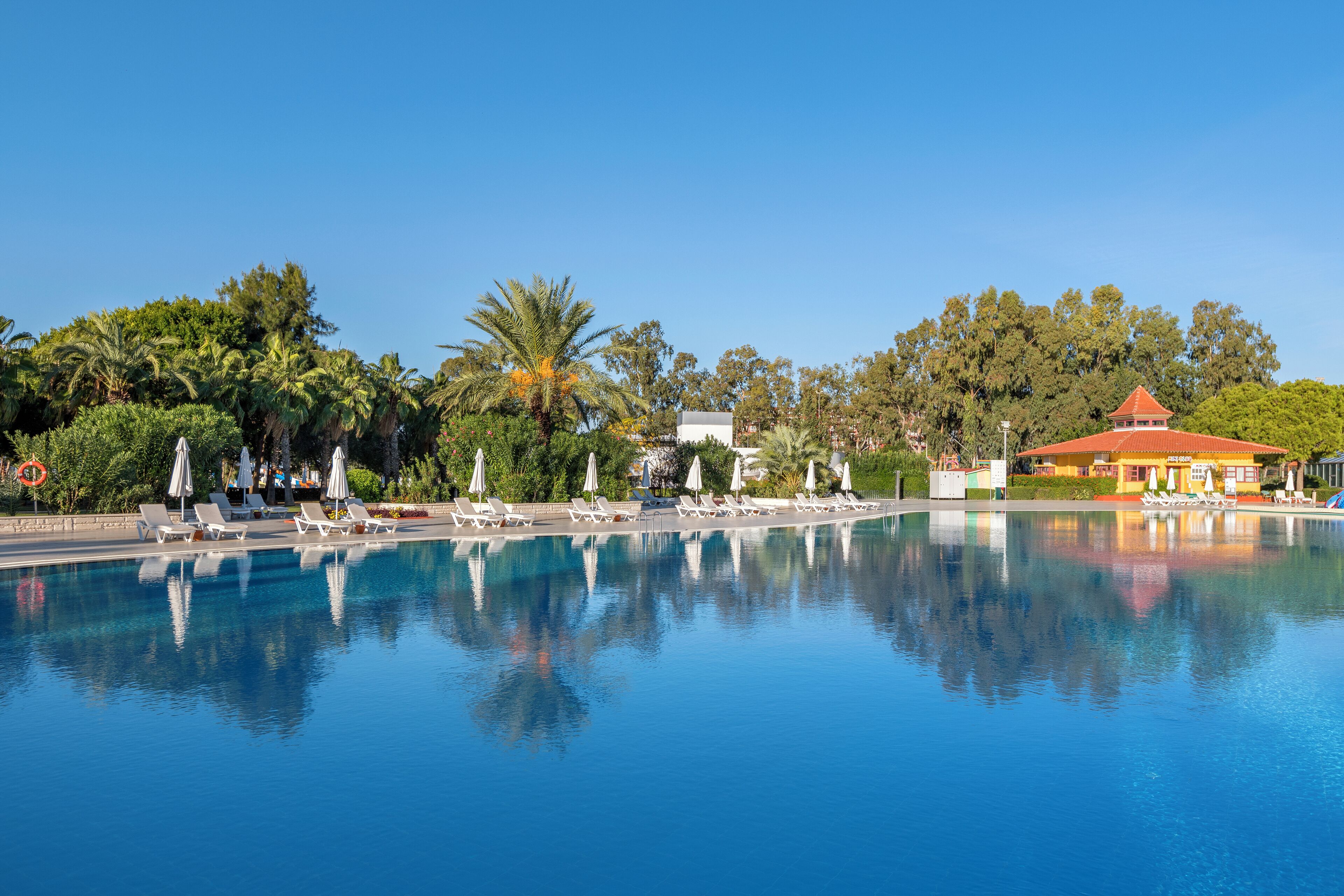 A clear swimming pool with lounge chairs and umbrellas, surrounded by trees and a building with a red roof.