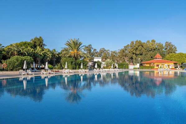 A clear swimming pool with lounge chairs and umbrellas, surrounded by trees and a building with a red roof. - Green Max Hotel (Serik)