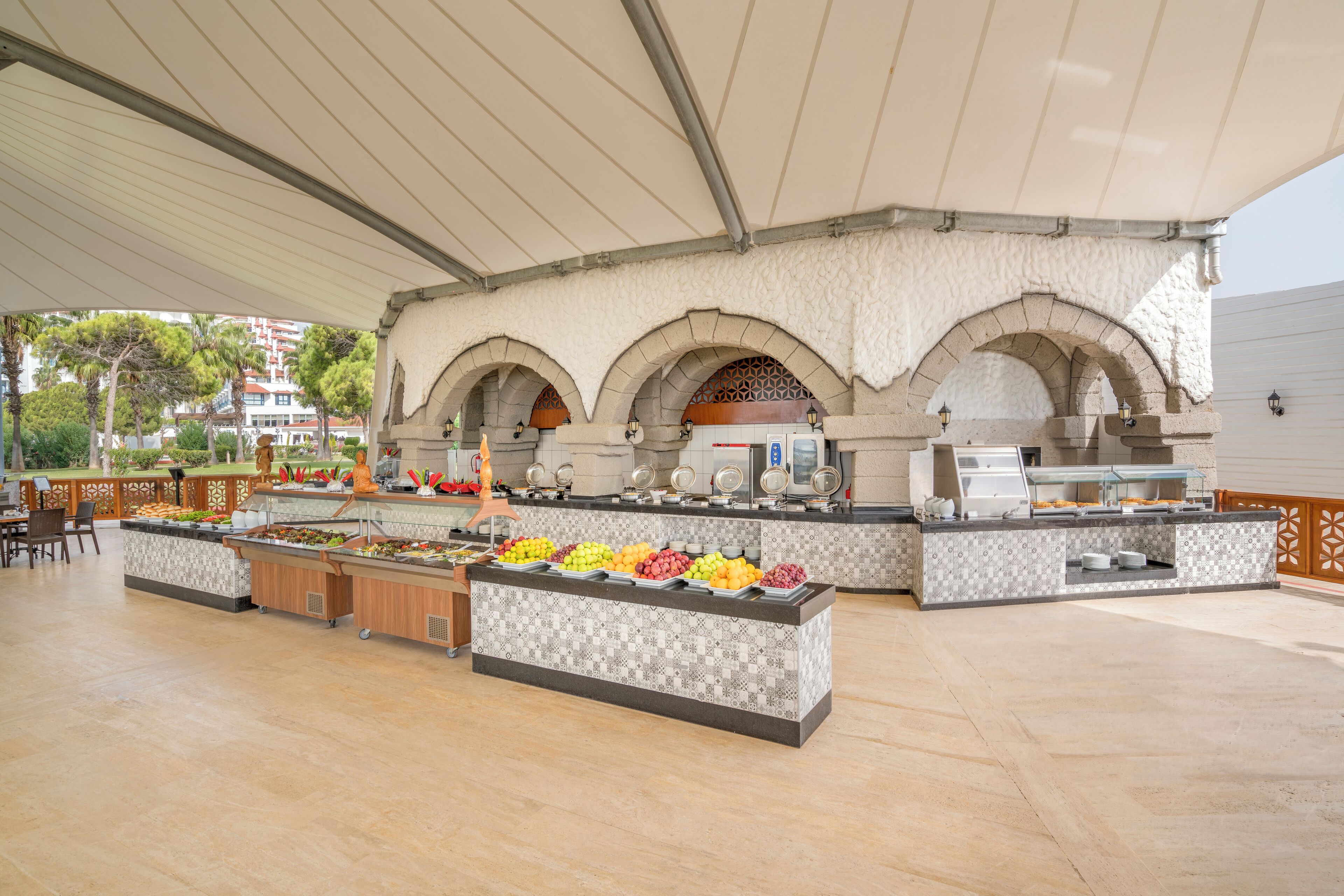 Zona de buffet al aire libre con suelo de baldosas, área de estar cubierta y un mostrador de buffet con diversas opciones de comida.