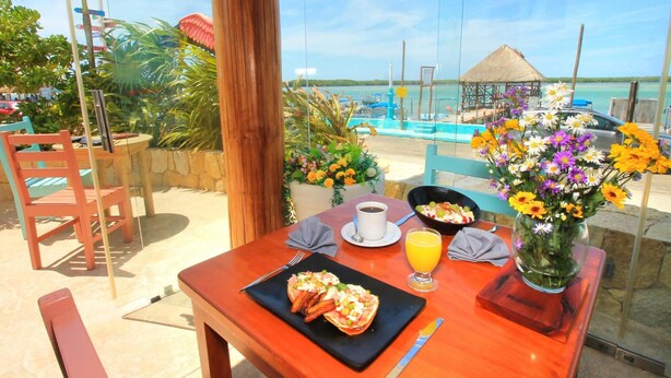 Een houten tafel met een ontbijtje, inclusief een bord eten, een kop koffie en een glas sinaasappelsap, met uitzicht op een strand met een rieten dak en een helderblauwe lucht.