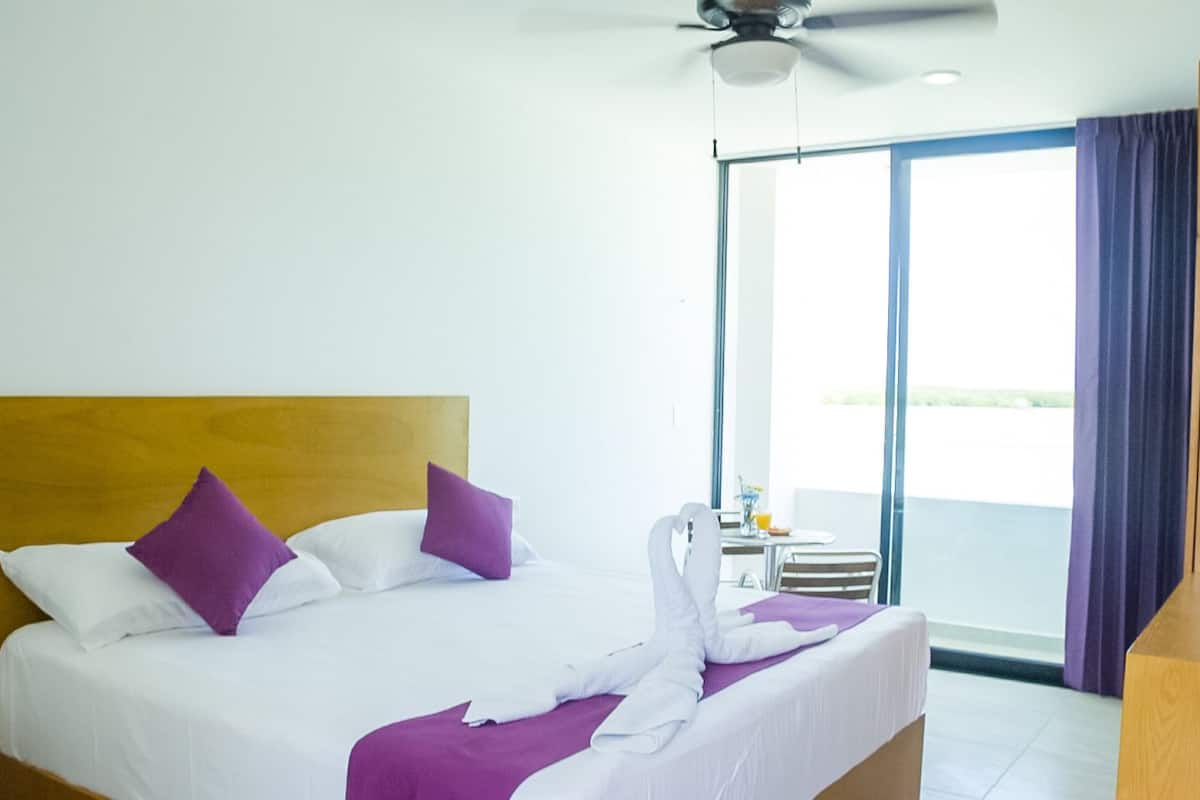 Habitación de hotel con cama, ventilador de techo, ventana con cortinas y vistas al exterior.