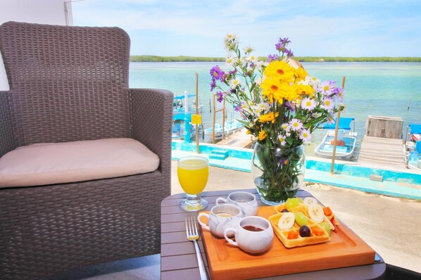 A wicker chair with a cushion, a table with a tray of fruit, a glass of orange juice, and a vase of flowers.