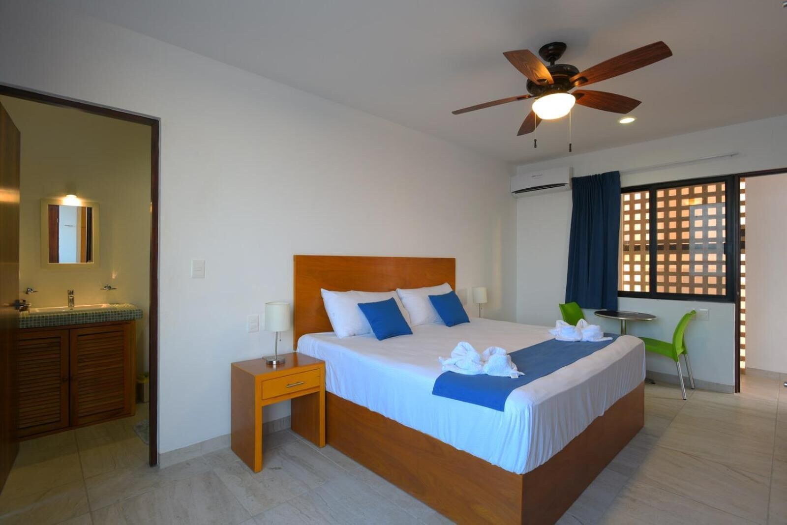 A hotel room with a wooden bed, a nightstand, a ceiling fan, a window with a view, and a bathroom with a sink and mirror.