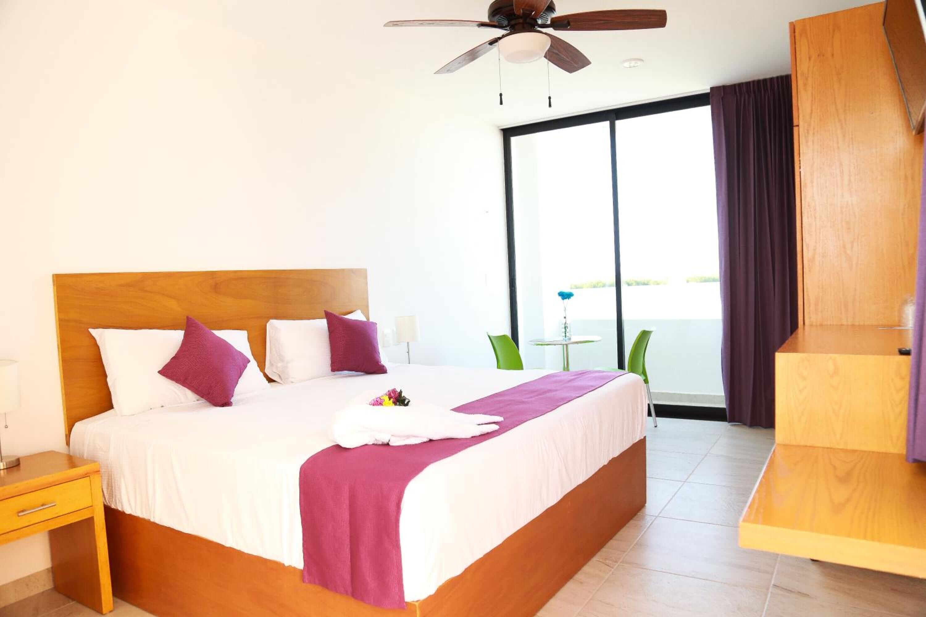 Habitación de hotel con una cama grande, ventilador de techo, vista al mar a través de una puerta corrediza de vidrio y una pequeña mesa con sillas.