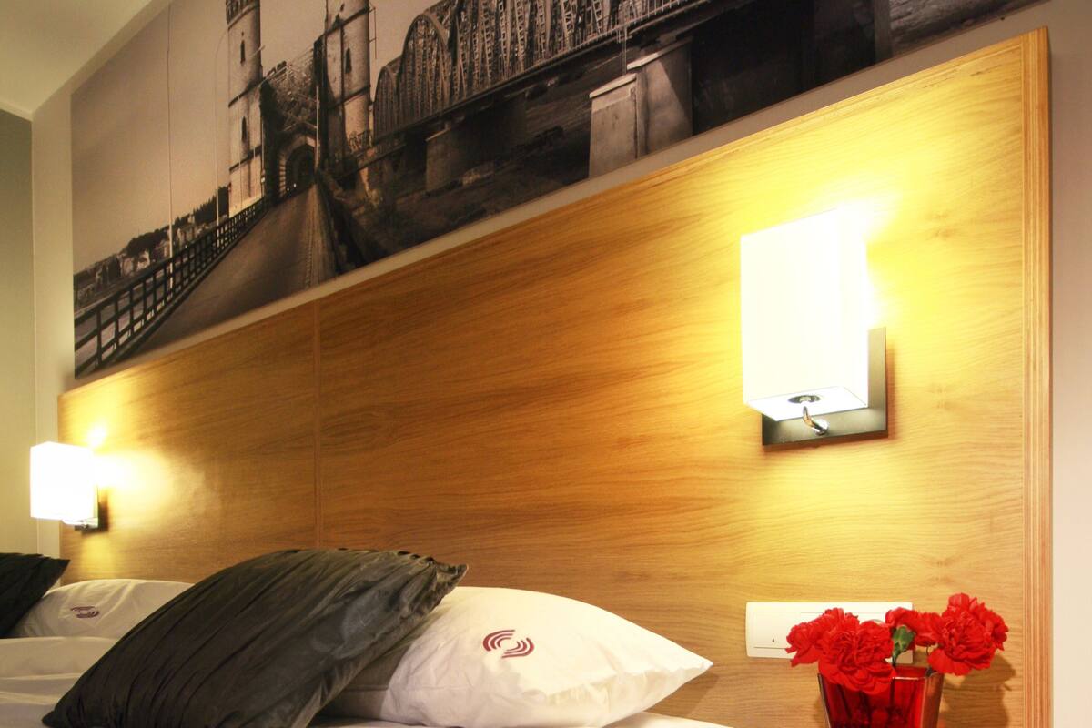 A hotel room with a wooden headboard, a bed with white bedding, a nightstand with a vase of red flowers, and a wall-mounted light fixture.