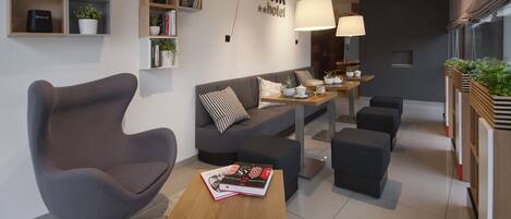 A modern hotel lobby with a grey sofa, black ottomans, a wooden coffee table, and a bookshelf with books and plants.