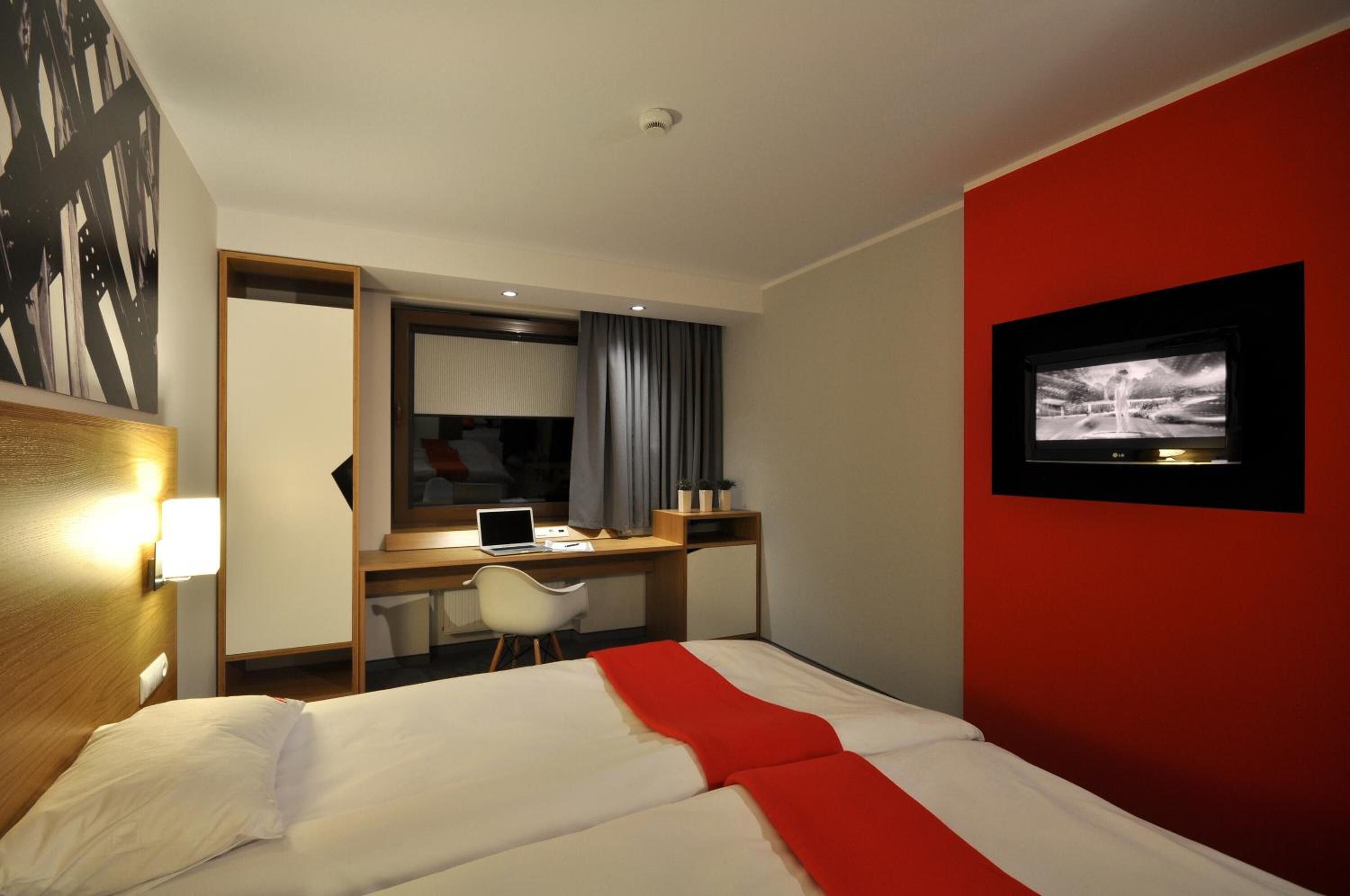 Una habitación de hotel con una cama, un escritorio con una computadora portátil, un televisor en la pared y una pared de acento roja.