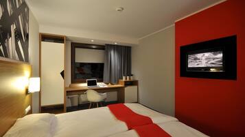 Una habitación de hotel con una cama, un escritorio con una computadora portátil, un televisor en la pared y una pared de acento roja.