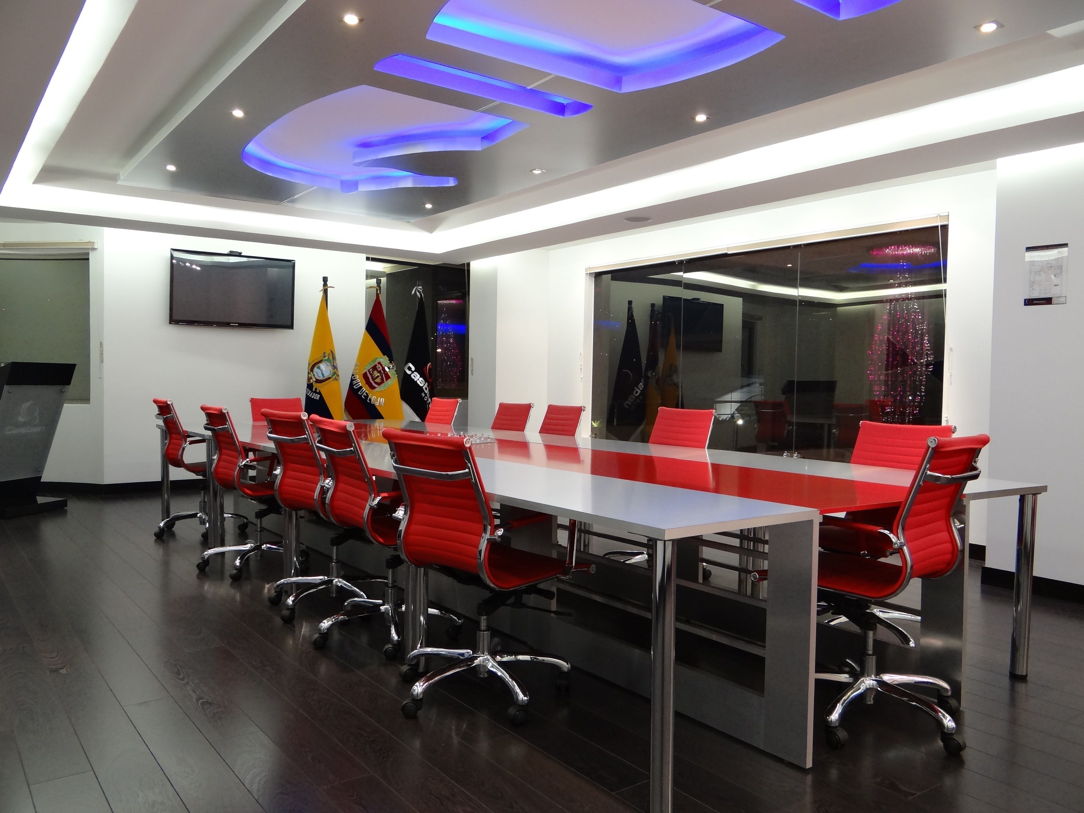 Uma sala de conferências com uma mesa comprida, cadeiras vermelhas e bandeiras na parede.