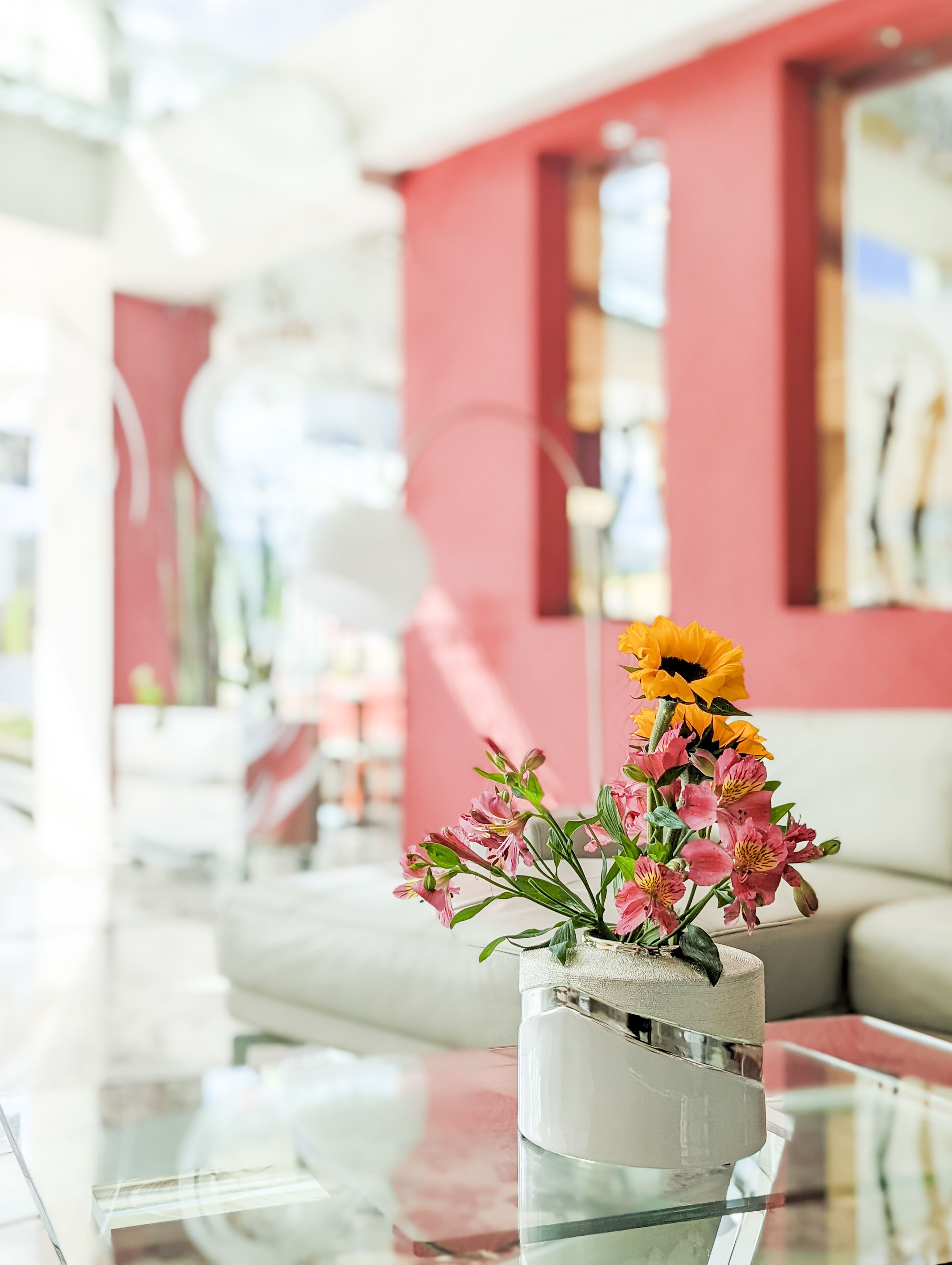 A modern living room with a glass coffee table, a vase with flowers, and a red wall.