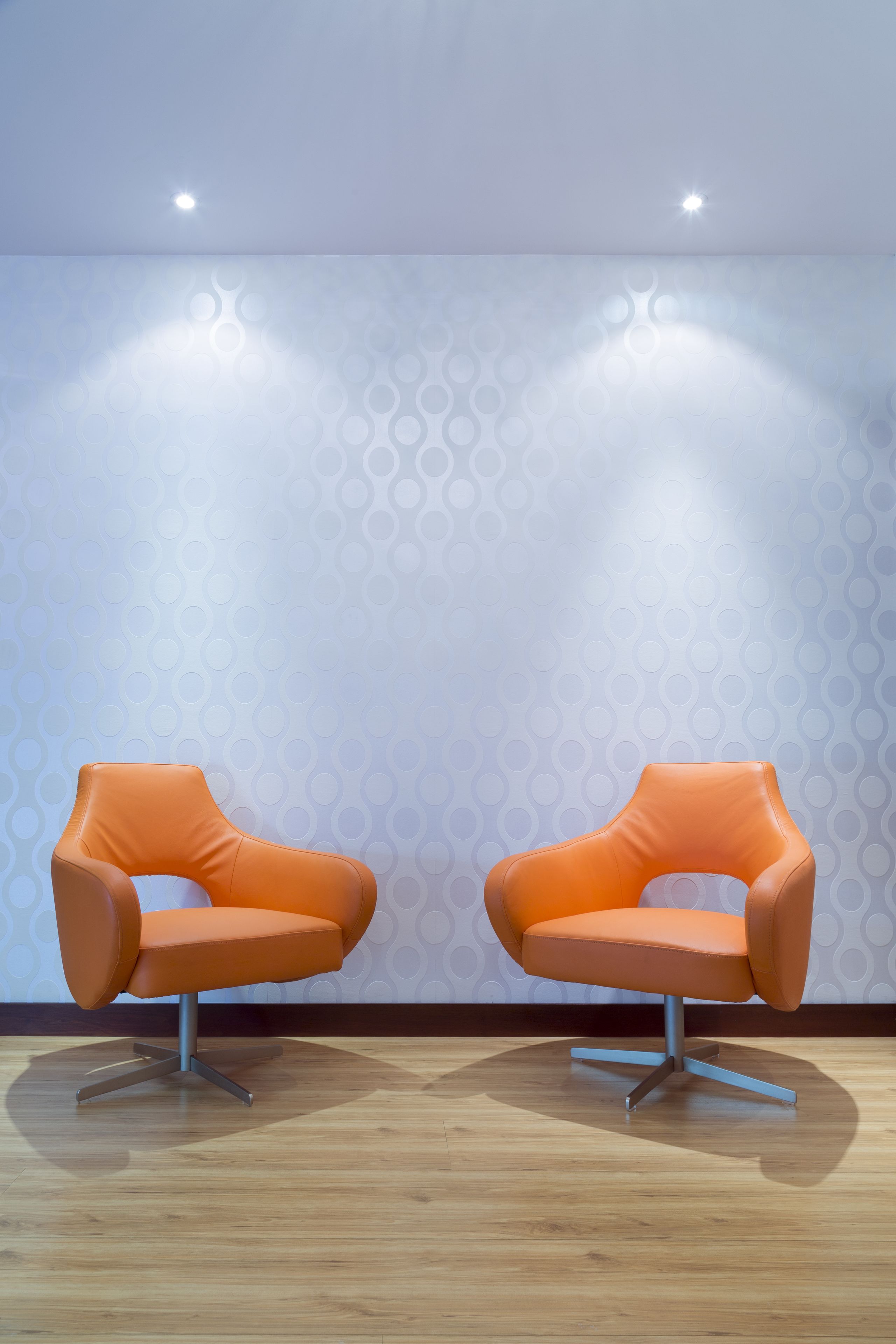 Two orange chairs with metal legs on a wooden floor, against a wall with a geometric pattern.