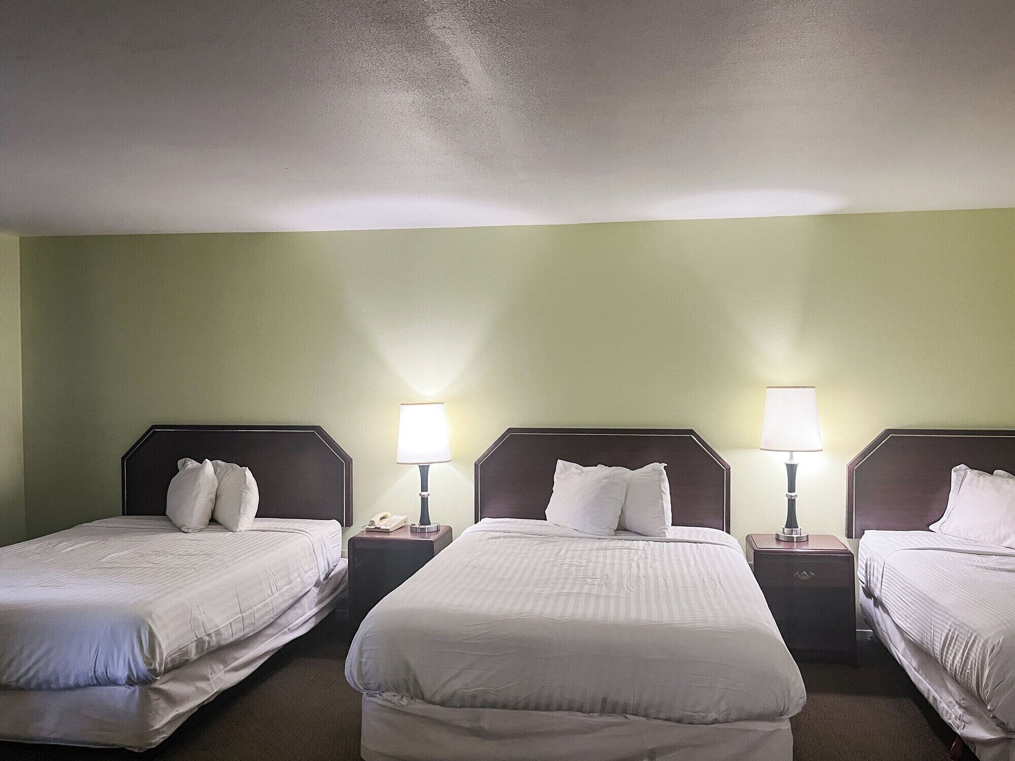 Habitación de hotel con tres camas individuales, cada una con una mesita de noche y una lámpara. Las paredes son de color verde claro y el techo es blanco.