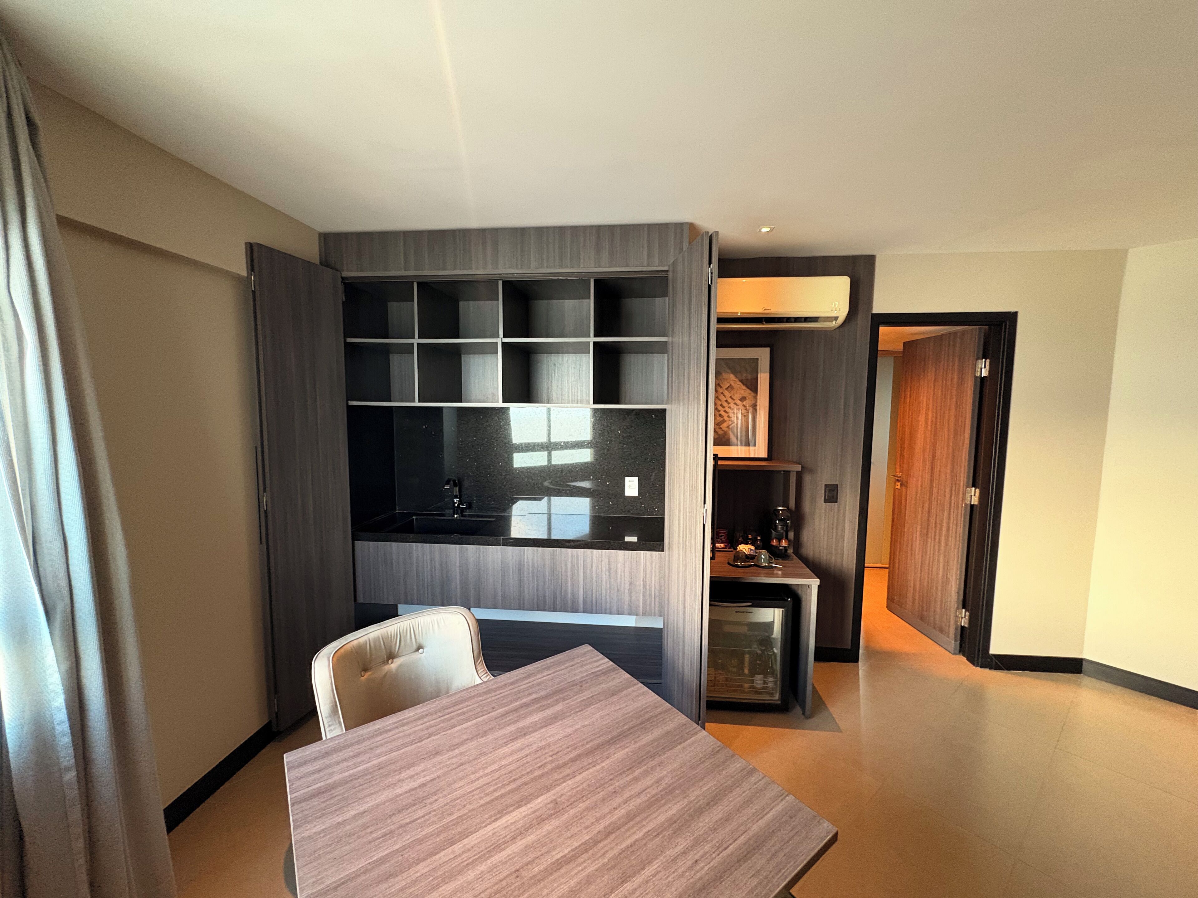 A modern hotel room with a wooden dining table, a built-in kitchen area, and a cozy seating arrangement.