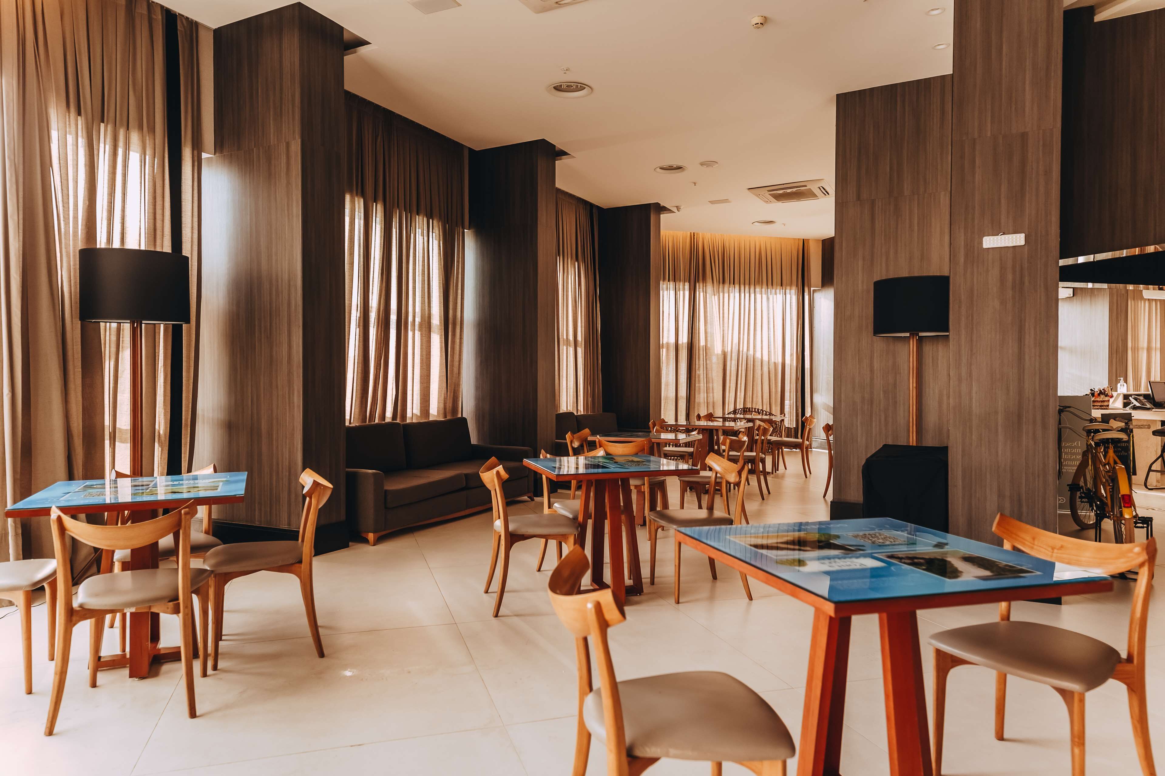 Uma sala de jantar moderna com mesas e cadeiras de madeira, grandes janelas com cortinas e uma bicicleta visível ao fundo.