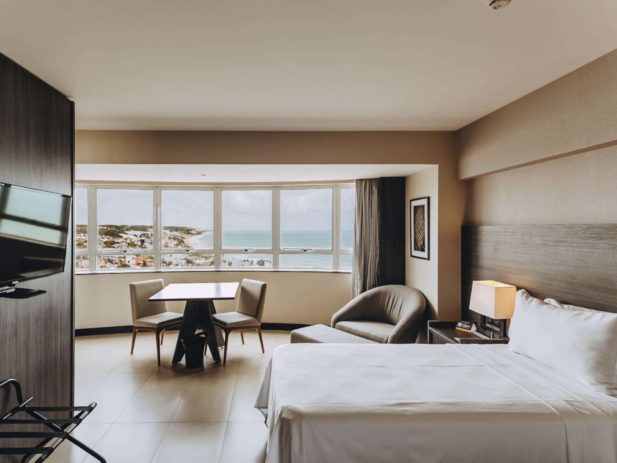 Habitación de hotel con una cama, un escritorio, dos sillas, un televisor, una ventana con vista a la playa y una lámpara.