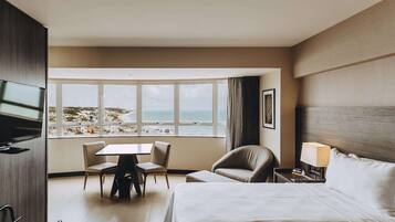 Habitación de hotel con una cama, un escritorio, dos sillas, un televisor, una ventana con vista a la playa y una lámpara.