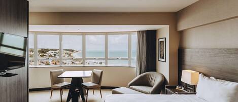 Habitación de hotel con una cama, un escritorio, dos sillas, un televisor, una ventana con vista a la playa y una lámpara.