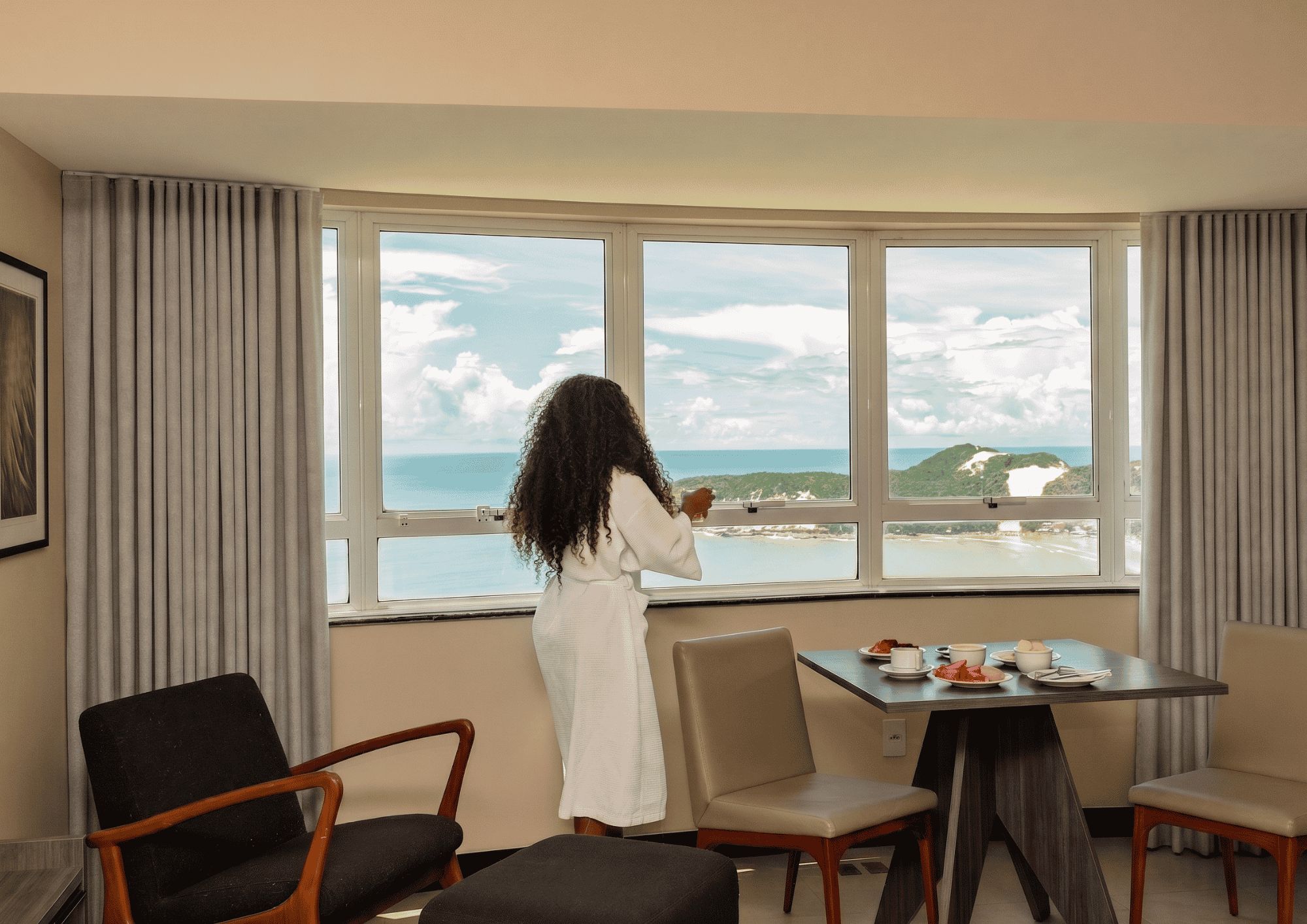 A room with a large window offering a view of the sea and hills, a dining table with plates and cups, and two chairs.