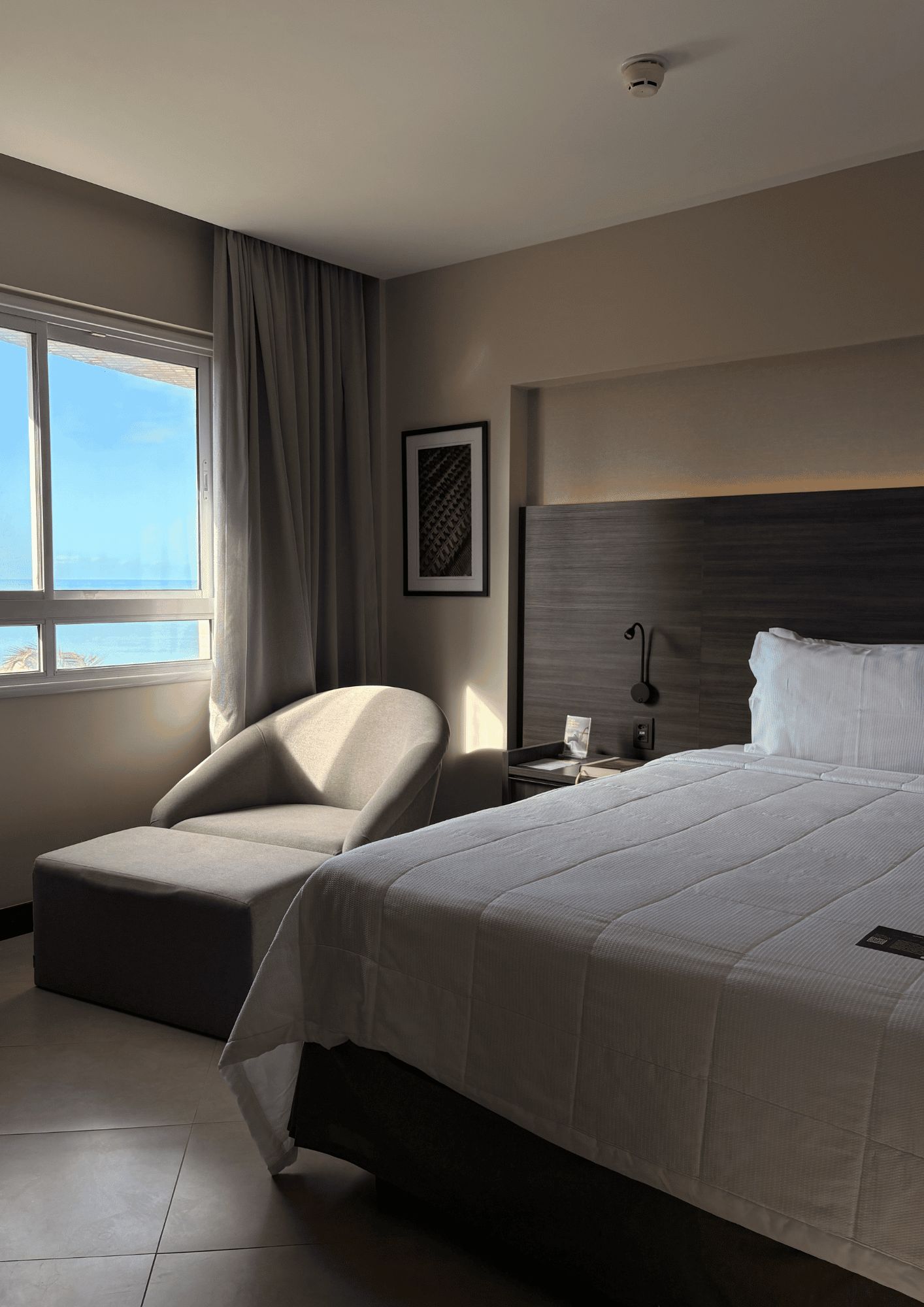 A hotel room with a bed, a chair, a nightstand, and a window with curtains.