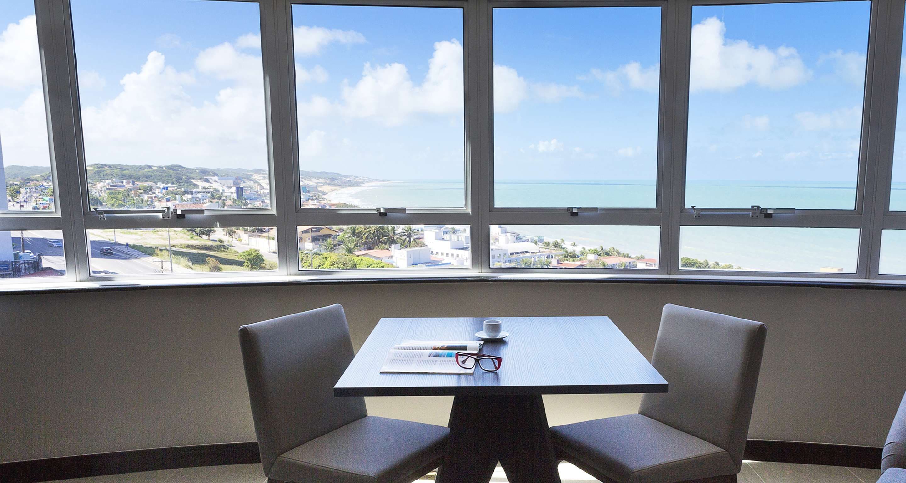 A room with a table and two chairs, a window with a view of a beach and cityscape.