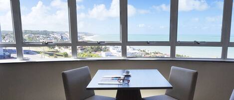 A room with a table and two chairs, a window with a view of a beach and cityscape.