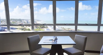 Quarto com mesa, duas cadeiras e janela com vista para a praia e paisagem urbana.