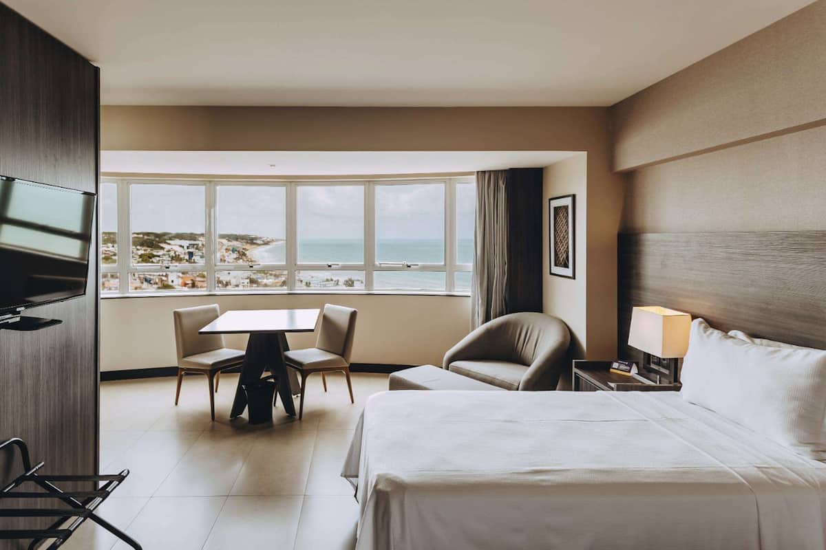 Ein Hotelzimmer mit Bett, Schreibtisch, zwei Stühlen, Fernseher, Fenster mit Meerblick und Lampe.