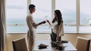 A couple in a room with a large window overlooking a body of water, a bottle of champagne, and a table with a cushion and a plate of food. - Golden Tulip Natal Ponta Negra (Natal)