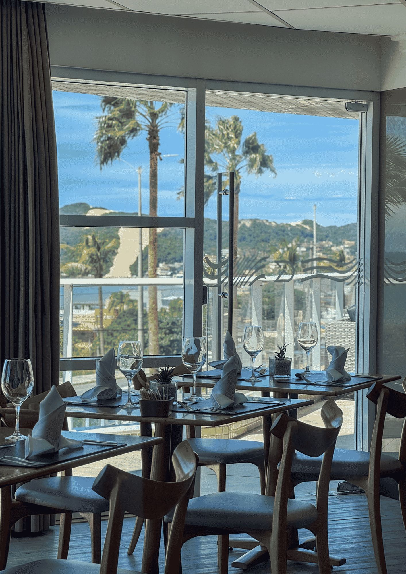 A dining area with tables set for two, overlooking a scenic view of palm trees and hills.