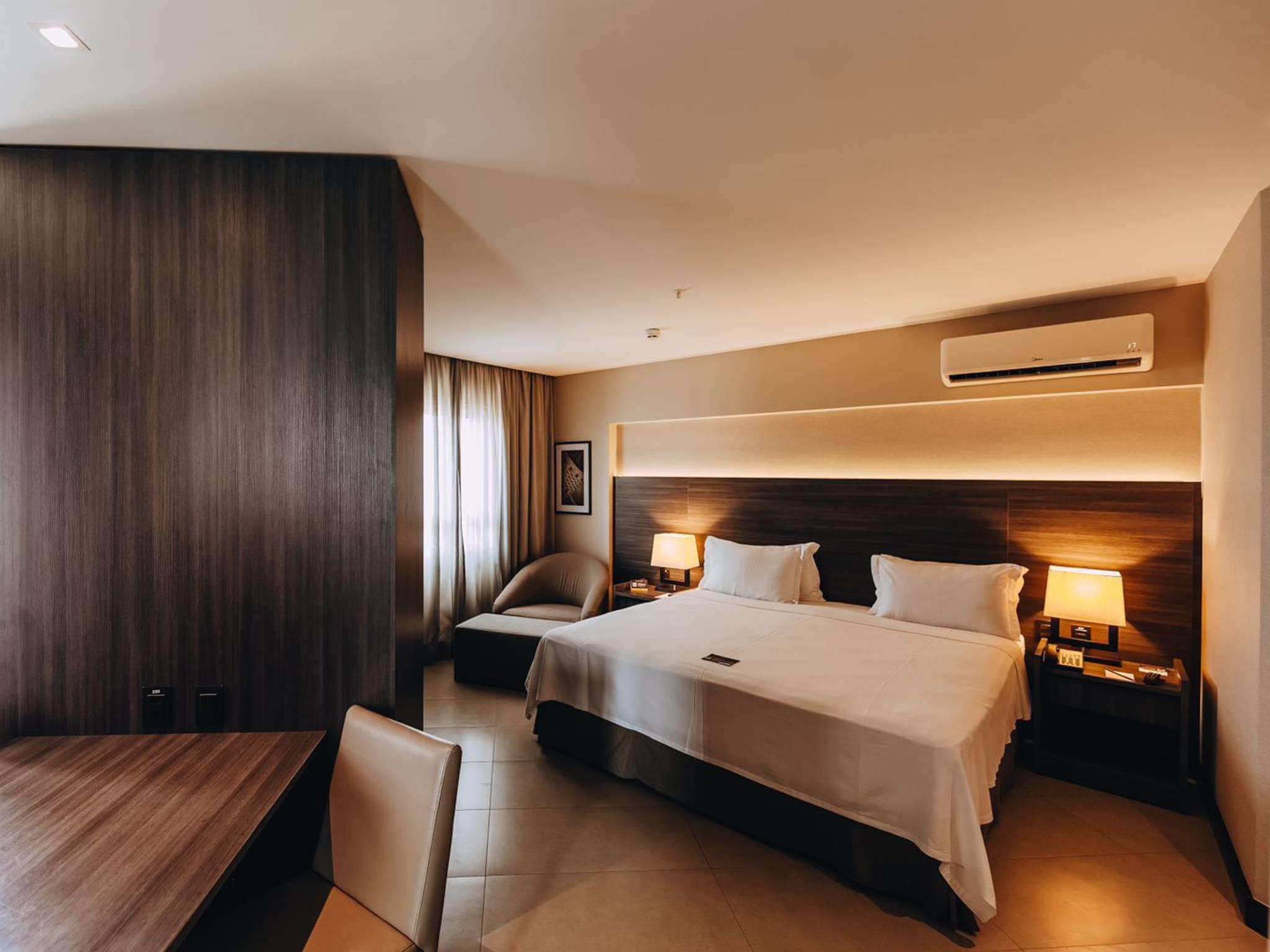 Habitación de hotel con una cama grande, un escritorio, una silla y un sofá. Hay un equipo de aire acondicionado en la pared y un cuadro colgado.