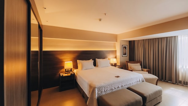 A hotel room with a large bed, two bedside tables with lamps, a wooden headboard, a sofa, and a large window with curtains. - Golden Tulip Natal Ponta Negra (Natal)