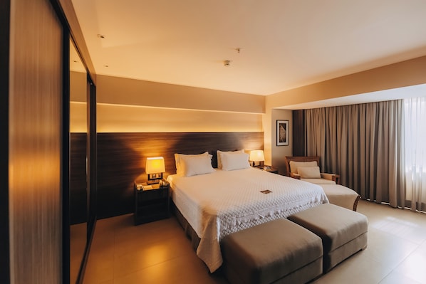 A hotel room with a large bed, two bedside tables with lamps, a wooden headboard, a sofa, and a large window with curtains.