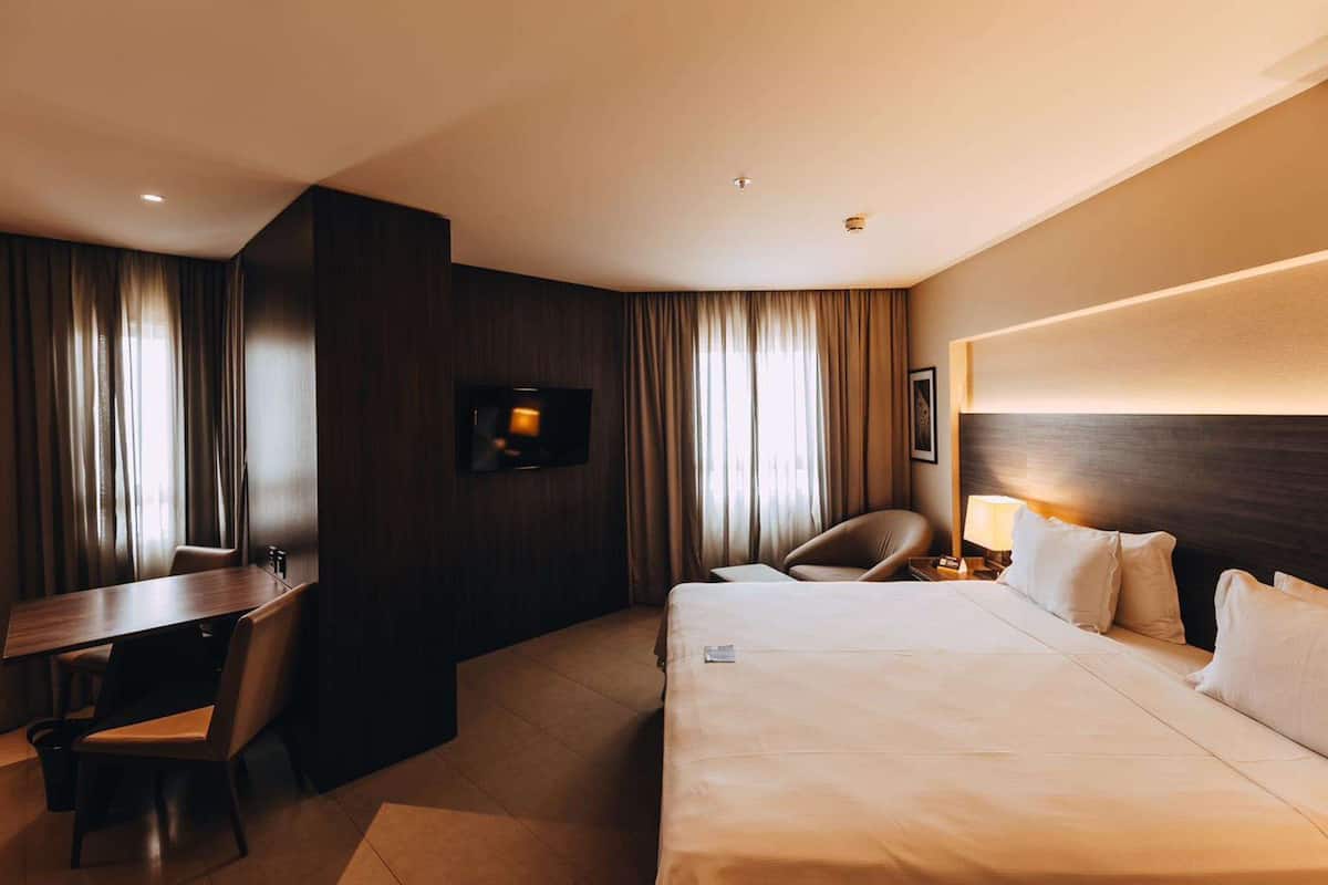 Ein Hotelzimmer mit einem großen Bett, einem Schreibtisch, einem Sessel, einem an der Wand befestigten Fernseher und einer Lampe auf dem Nachttisch.
