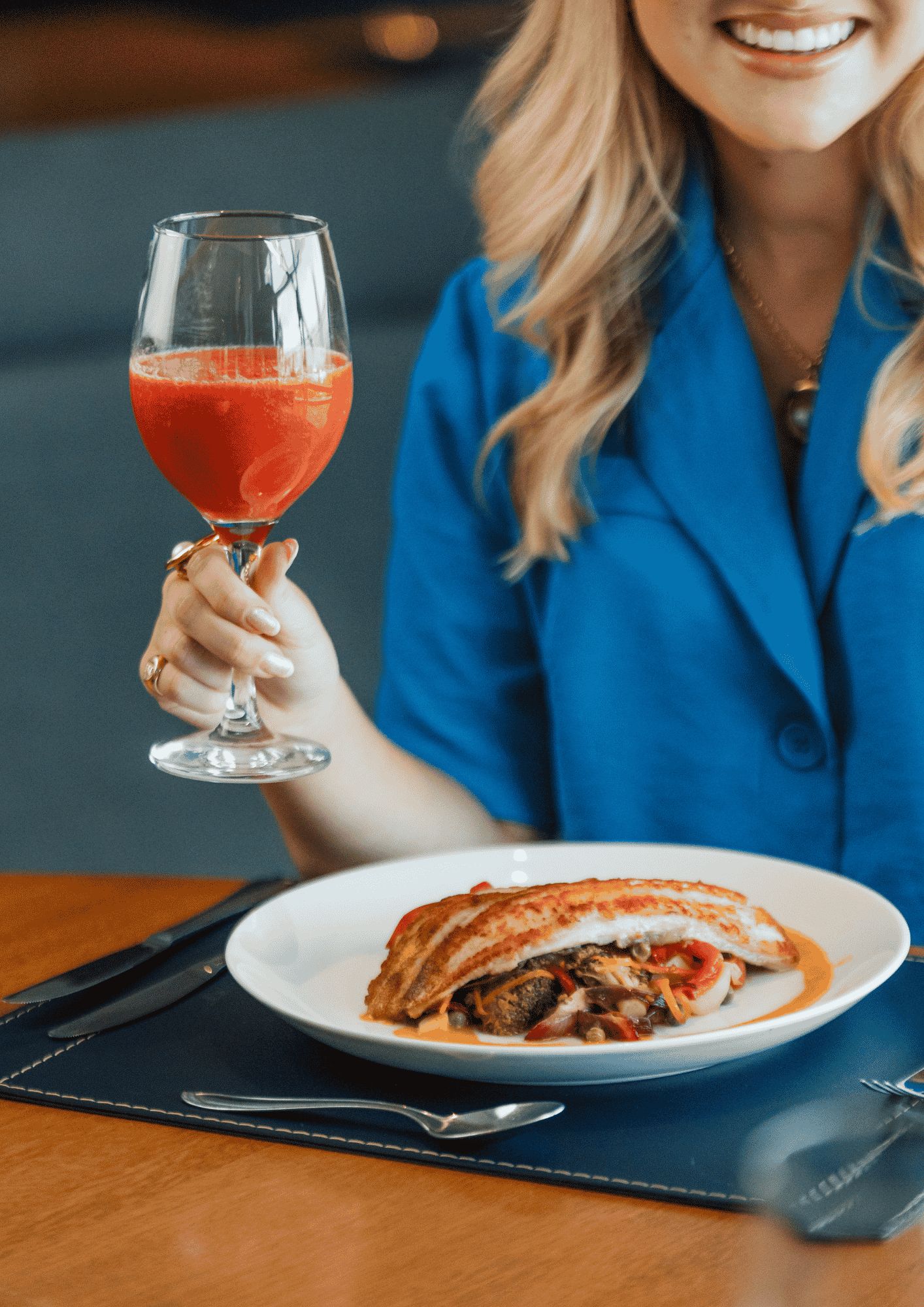 A person holding a glass of red drink, sitting at a table with a white plate of food.