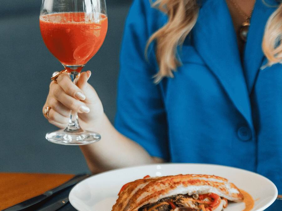 Una persona sosteniendo un vaso de bebida roja, sentada a una mesa con un plato blanco de comida.